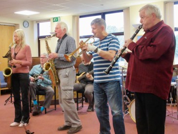 Four people playing brass/woodwind instruments, with a guitarist, bass guitarist and drummer in the background.